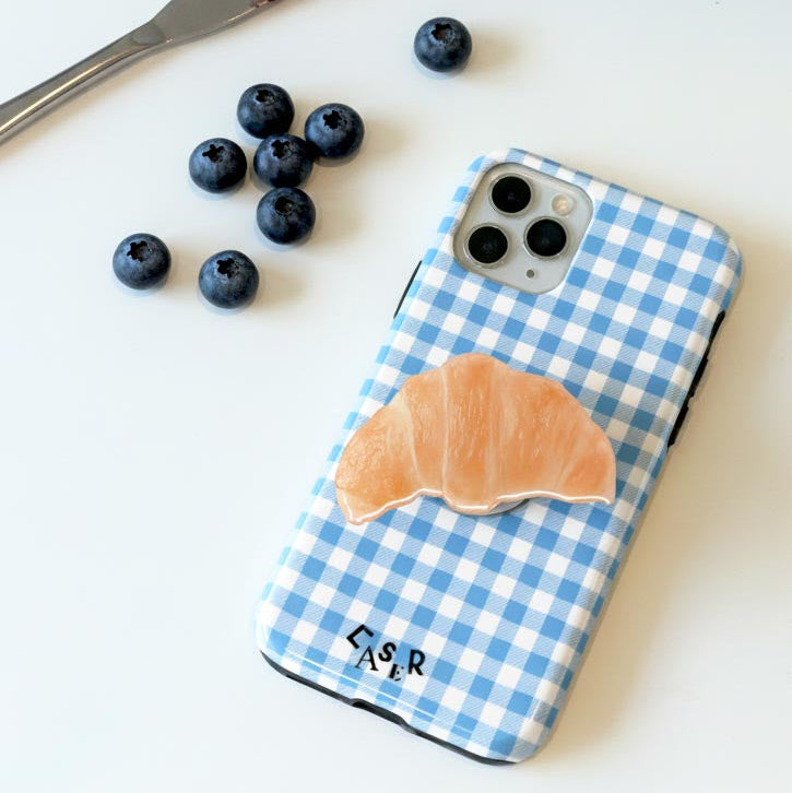 Phone case with blue checkered pattern and croissant phone grip on a white surface with berries and a knife.